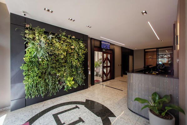 Un vestíbulo con mostrador de recepción, una pared con jardín vertical y una puerta con la inscripción 'SAIDA'.