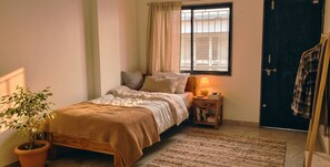 A bedroom with a bed, bedside table, lamp, potted plant, and a view of buildings through the window. - Fantastic 3-bedroom apartment in Nashik perfect for your stay  and relaxation. (Nashik)