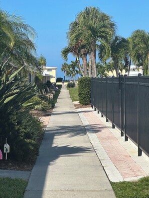 A paved pathway flanked by palm trees and a metal fence, leading to a building with a yellow facade. - Charming Coastal Studio | Steps to Private Beach | Patio | Casa Mar Resort (Sarasota)