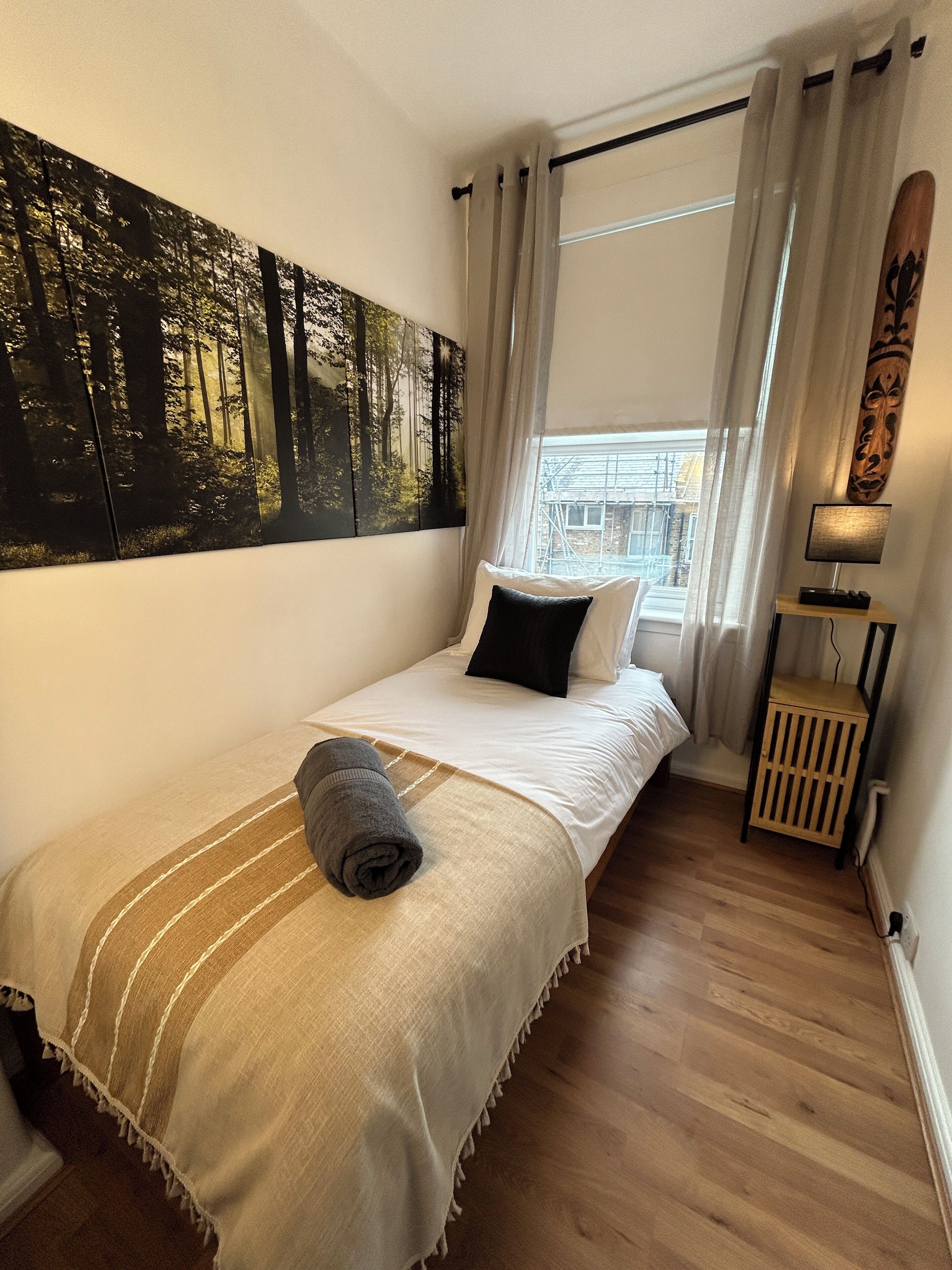 A bedroom with a bed, a large forest-themed painting, a window with curtains, a bedside lamp, and a wooden nightstand.