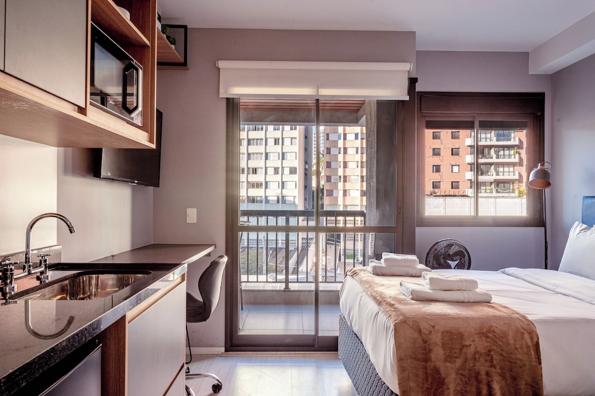 A modern hotel room with a kitchenette, a large window with a city view, and a bed with a brown blanket.