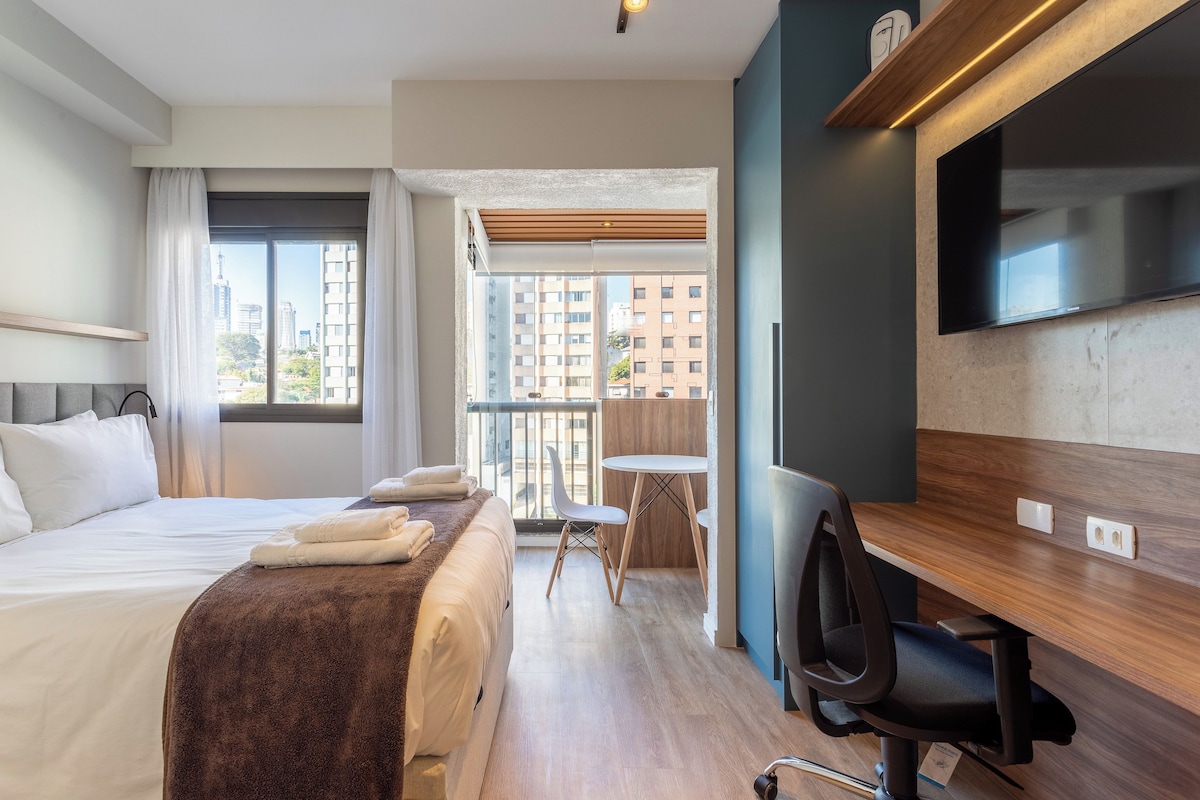 A hotel room with a bed, a desk, a chair, a TV, and a balcony with a view of the city.