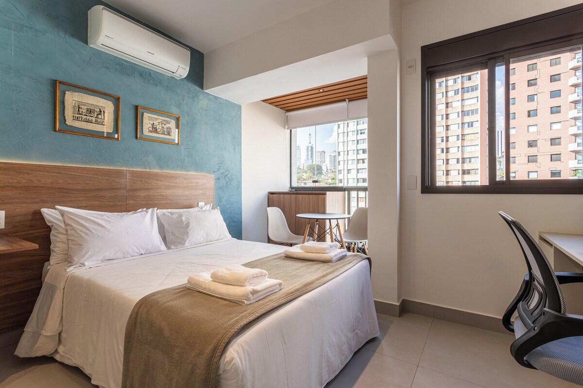 A hotel room with a bed, a desk, a chair, a window with a city view, and an air conditioning unit.