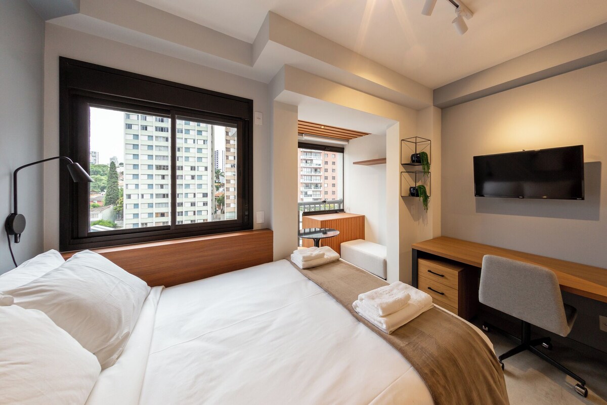 A modern hotel room with a large bed, a desk, a chair, a TV, and a window with a city view.