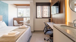 A hotel room with a bed, a desk, a chair, a TV, and a window with a view of buildings. - Update by Bhomy (São Paulo)
