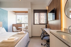 A hotel room with a bed, a desk, a chair, a TV, and a window with a view of buildings. - Update by Bhomy (São Paulo)