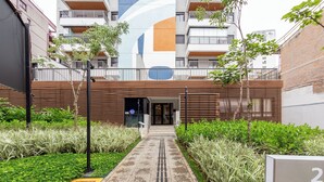 A modern building with a large vertical mural, a gated entrance, and a pathway leading to the building. - Update by Bhomy (São Paulo)