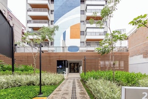 A modern building with a large vertical mural, a gated entrance, and a pathway leading to the building. - Update by Bhomy (São Paulo)