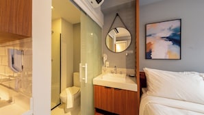 A modern bathroom with a glass shower enclosure, a toilet, a sink with a mirror, and a bed with white bedding. - Update by Bhomy (São Paulo)