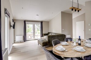A modern dining area with a round wooden table set for four, a sofa, and a view through a glass door to an outdoor patio. - Villa Riant 58 | Ruim vakantiehuis, hondvriendelijk | Betuwe (Zennewijnen)