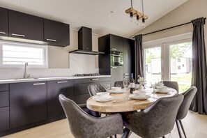 A modern kitchen with dark cabinetry, a central island, and a dining area with grey upholstered chairs. - Villa Riant 58 | Ruim vakantiehuis, hondvriendelijk | Betuwe (Zennewijnen)