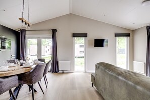 A modern living room with a sofa, a dining table set for four, and a television mounted on the wall. - Villa Riant 58 | Ruim vakantiehuis, hondvriendelijk | Betuwe (Zennewijnen)