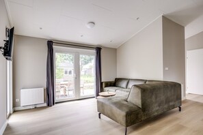 A modern living room with a sectional sofa, a flat-screen TV mounted on the wall, and a sliding glass door leading to an outdoor patio. - Villa Riant 58 | 6 persoons vakantiehuis (Zennewijnen)