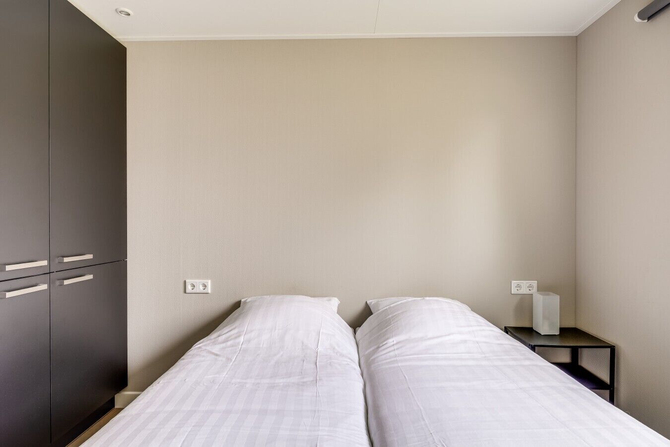 A bedroom with two single beds, a wardrobe, a bedside table with a lamp, and a power outlet on the wall.