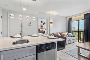 Een moderne keuken met een vaatwasser, een eethoek met een tafel en stoelen, een bank met kussens, en een groot raam met uitzicht.