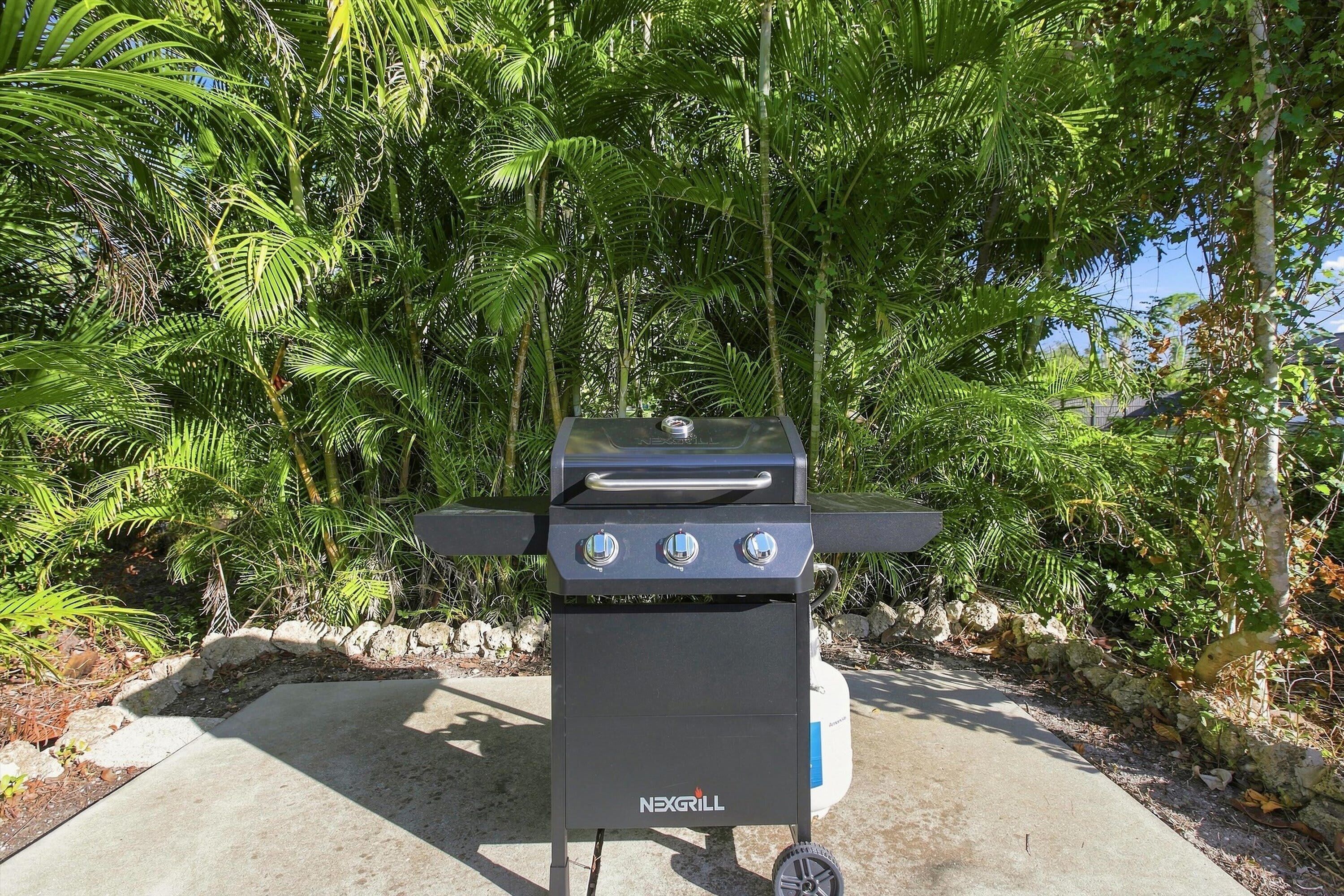 A black outdoor grill with three control knobs, set on a concrete surface surrounded by greenery.