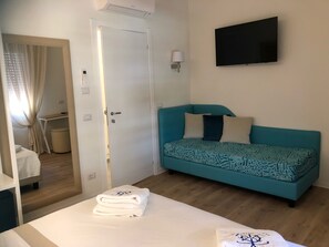 A room with a teal sofa, a bed with white linens, a TV mounted on the wall, and a door leading to another room.
