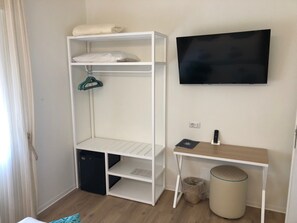 A room with a white shelving unit, a black TV mounted on the wall, a small wooden desk, and a trash can.