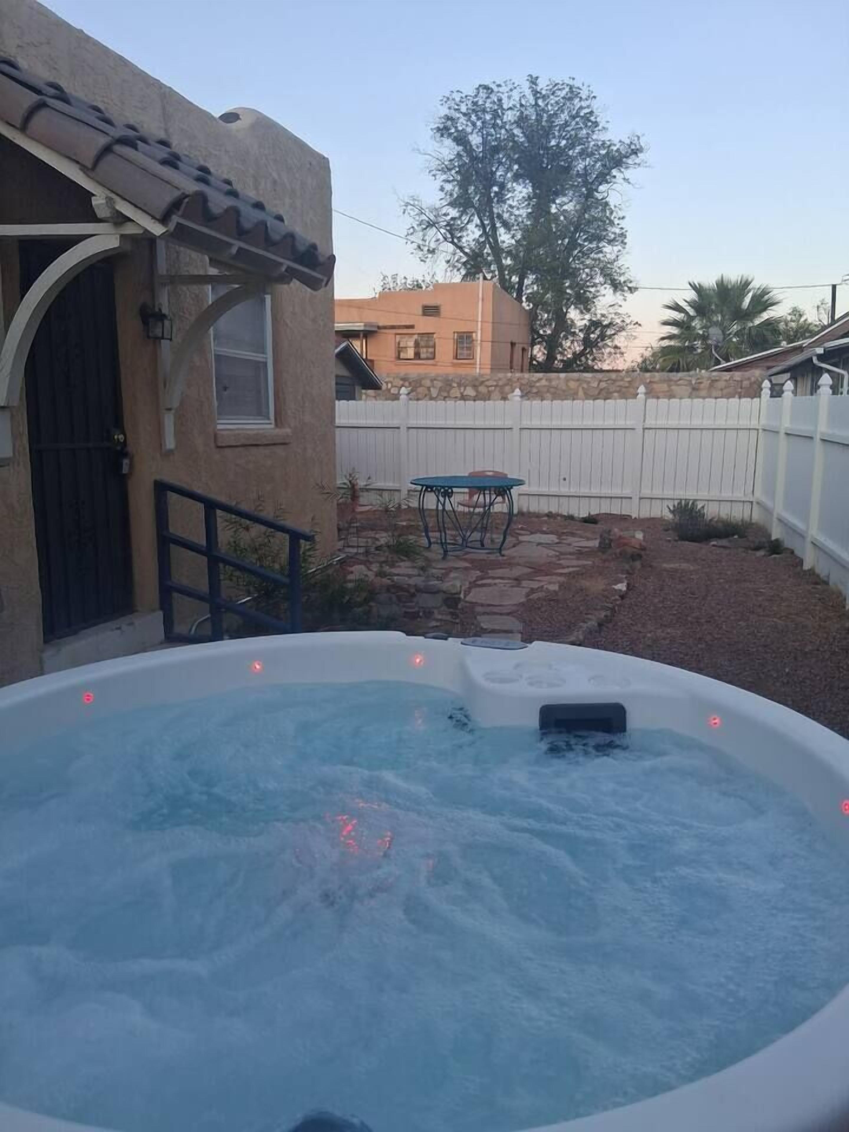 A hot tub in a backyard with a stone pathway, a wooden fence, and a building with a tiled roof.