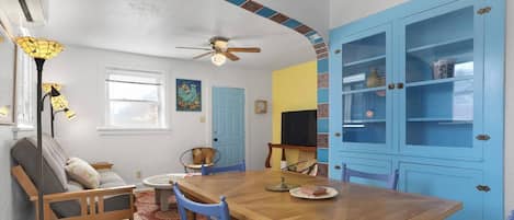 Un coin repas avec une table en bois, des chaises bleues, un ventilateur de plafond et un buffet bleu avec des portes en verre.
