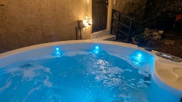 A hot tub with a person in it, surrounded by a stone wall and a covered area with string lights.