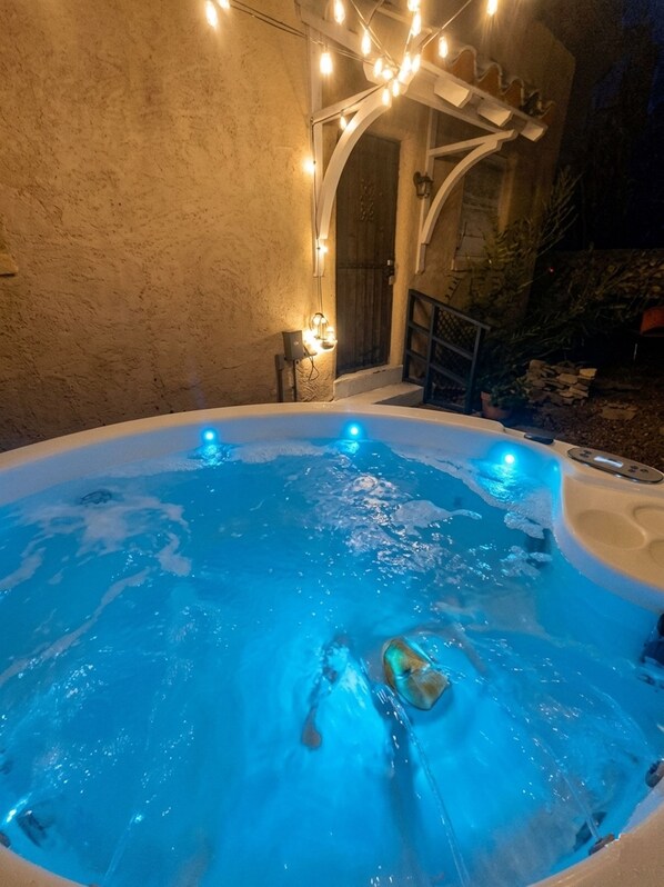 Un jacuzzi con una persona adentro, rodeado por un muro de piedra y un área techada con luces colgantes.