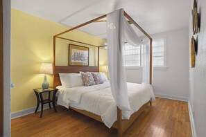 Una cama con dosel con ropa de cama blanca, una mesita de noche con una lámpara y un cuadro en la pared.