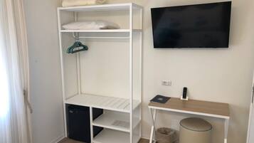 Ein Zimmer mit einer weißen Regaleinheit, einem an der Wand montierten schwarzen Fernseher, einem kleinen Holztisch und einem Mülleimer.