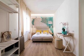 Una camera d'albergo con un letto, una scrivania, una sedia, un tavolino, un cestino e una mappa appesa al muro.