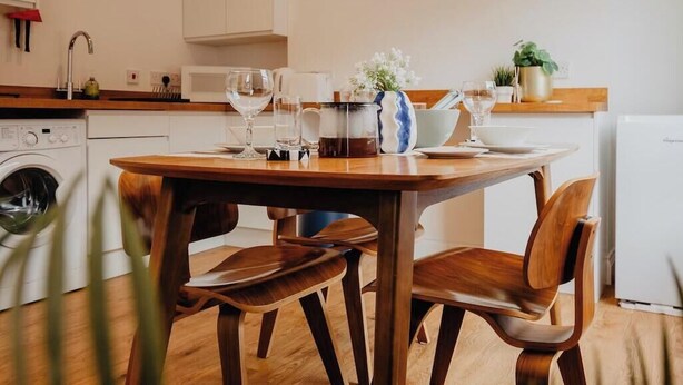 Un coin repas avec une table et des chaises en bois, une horloge au mur et une cuisine en arrière-plan.