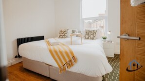 Une chambre à coucher avec un lit, une fenêtre, un radiateur, une table de chevet et une porte.
