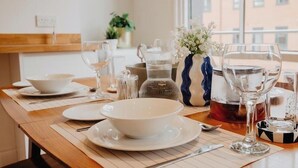 Une table à manger dressée avec de l’assiette, des bols et des couverts blancs, agrémentée d’un vase de fleurs et d’une théière posée sur la table.