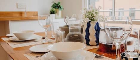 A dining table set with white plates, bowls, and cutlery, with a vase of flowers and a teapot on the table.