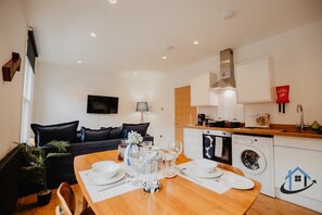 A modern living room with a sofa, a dining table set for four, a kitchen area with a washing machine, and a flat-screen TV mounted on the wall.