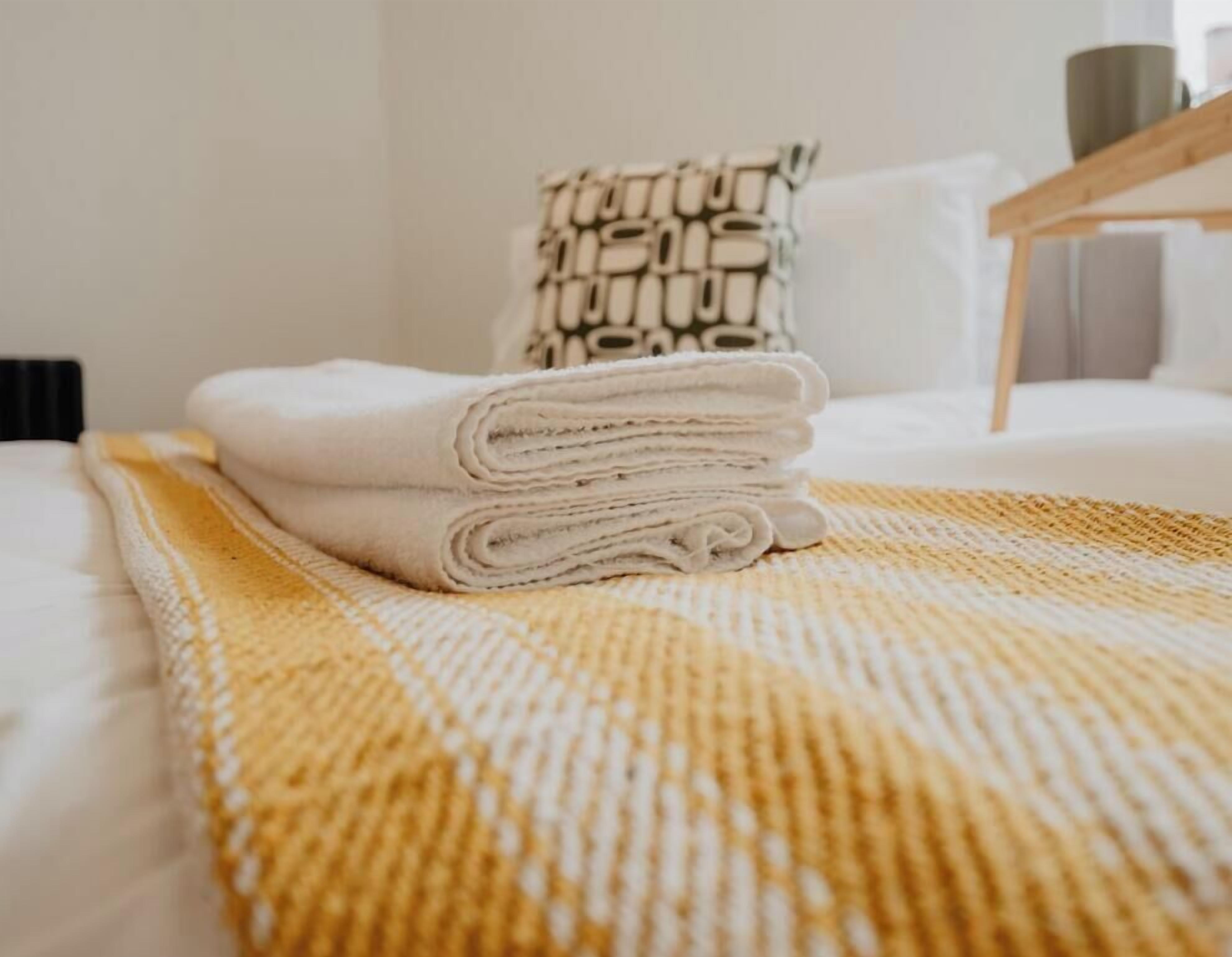 Una cama bien hecha con una toalla blanca doblada encima, un cojín estampado y una mesita de noche de madera.