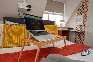 Una laptop sobre un atril de madera sobre una cama con almohadas amarillas y blancas, una manta roja y una alfombra roja.