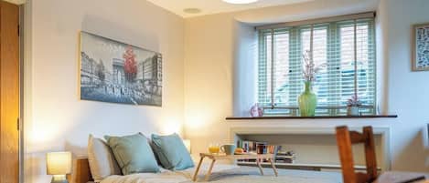 Une chambre à coucher comprenant un lit, une table de chevet, une lampe, une fenêtre avec des stores, des étagères et un tableau accroché au mur.