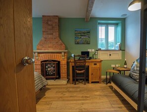 Een kamer met een schouw van baksteen, een houten bureau, een stoel, een bank en een raam met luxaflex.