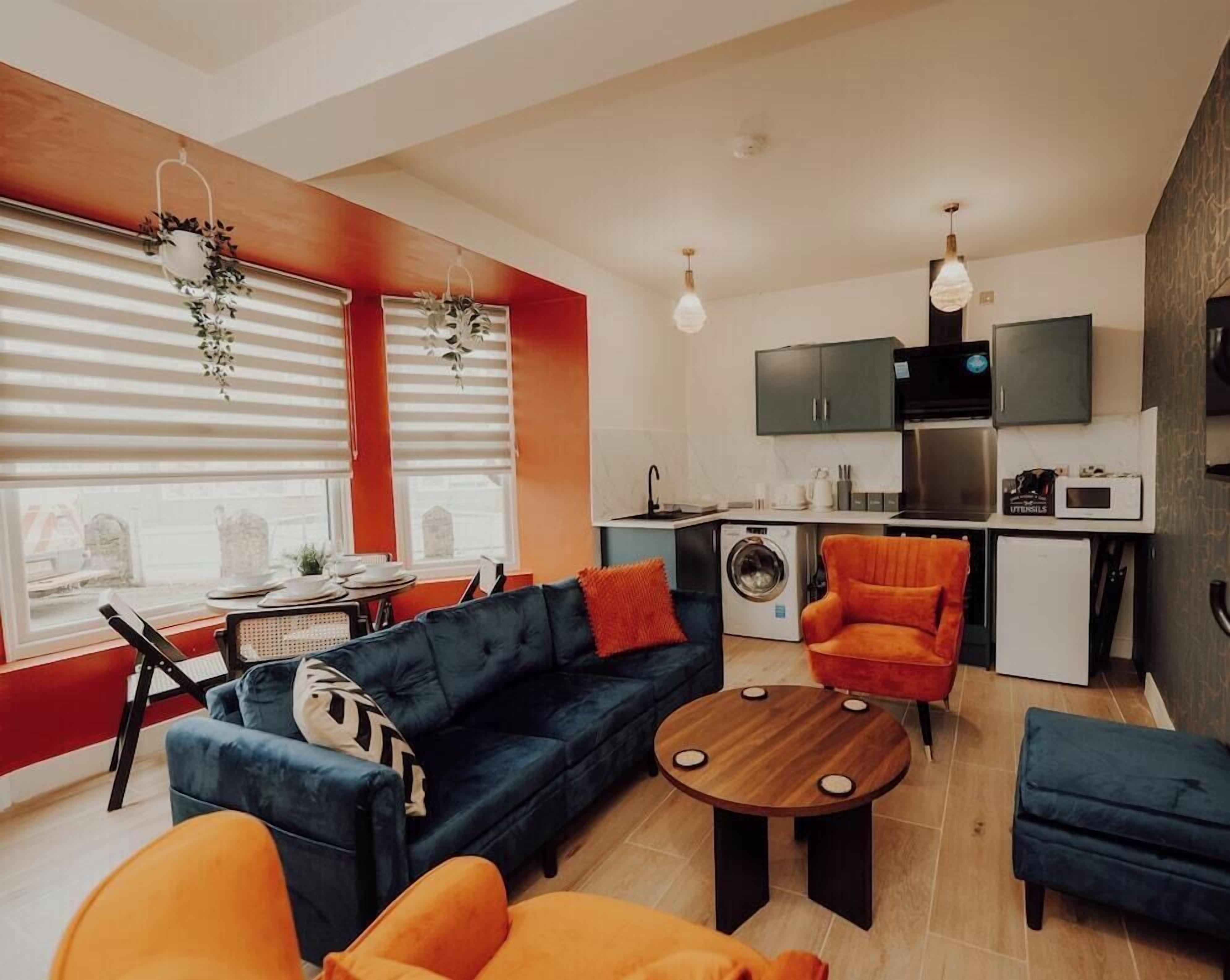 Ein modernes Wohnzimmer mit einer Couch, einem Sessel und einem Hocker, einer Küchenzeile mit Geräten und einem großen Fenster mit Jalousien.