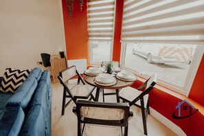 Ein Essbereich mit einem runden Tisch, gedeckt für vier Personen, umgeben von Korbstühlen, einem blauen Sofa und einem Fenster mit Jalousien.