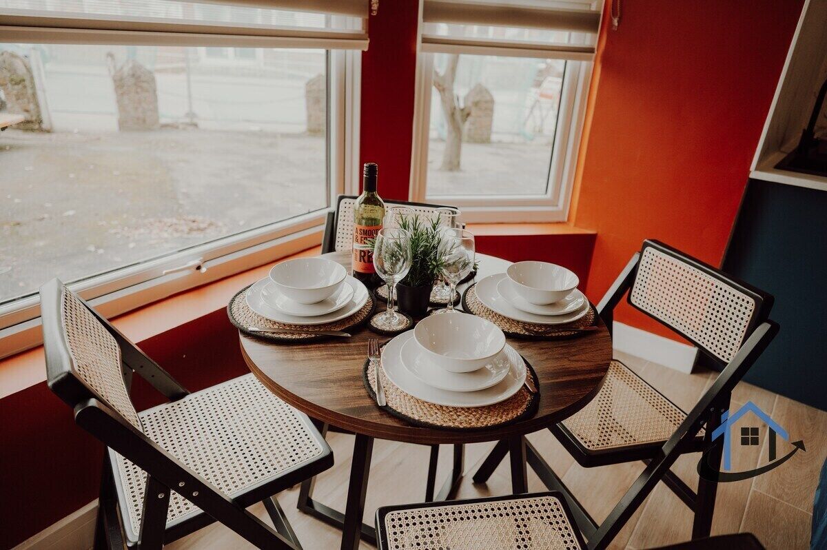 A round dining table with four white plates, two wine glasses, and a bottle of wine, set against a red wall with large windows.