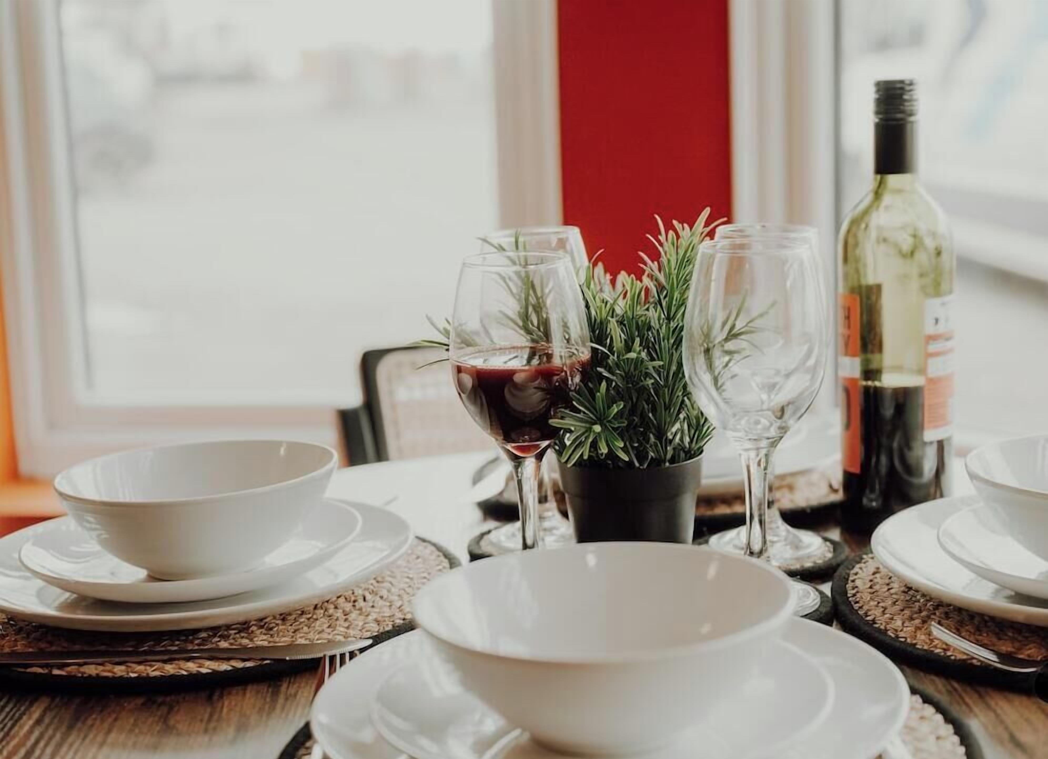 A dining table set with white plates, bowls, and wine glasses, with a bottle of wine and a potted plant in the background.