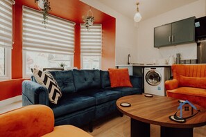 Een moderne woonkamer met een donkerblauwe bank, oranje fauteuils, een houten salontafel en een keuken met een wasmachine.