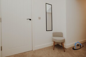 Een witte deur met een zwarte handgreep, een kleine stoel met houten poten en een spiegel aan de muur.