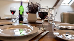 A dining table set with plates, cutlery, and wine glasses, with a bottle of wine and a vase of dried flowers in the background.