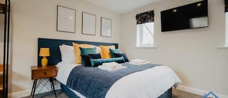 A bedroom with a bed, a nightstand, a lamp, framed certificates on the wall, a TV, and a window.