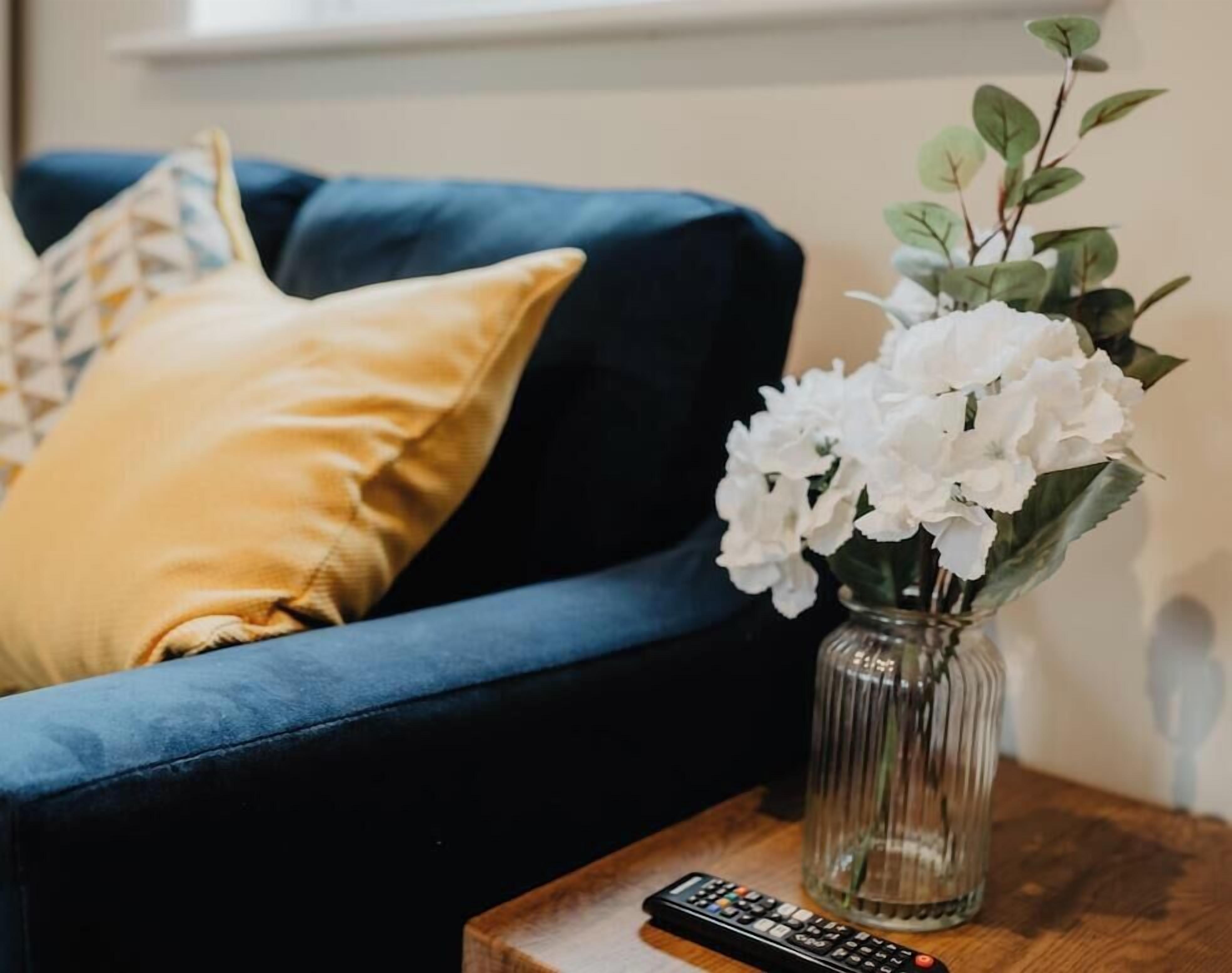 Un canapé bleu avec un coussin jaune, un vase avec des fleurs blanches et une télécommande sur une petite table en bois.