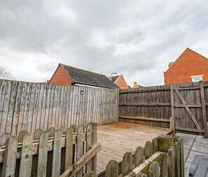 A wooden deck with a wooden fence, a brick building with a red roof, and a cloudy sky. - Ironside House 3 bed house, sleeps 8 parking (Thrapston)