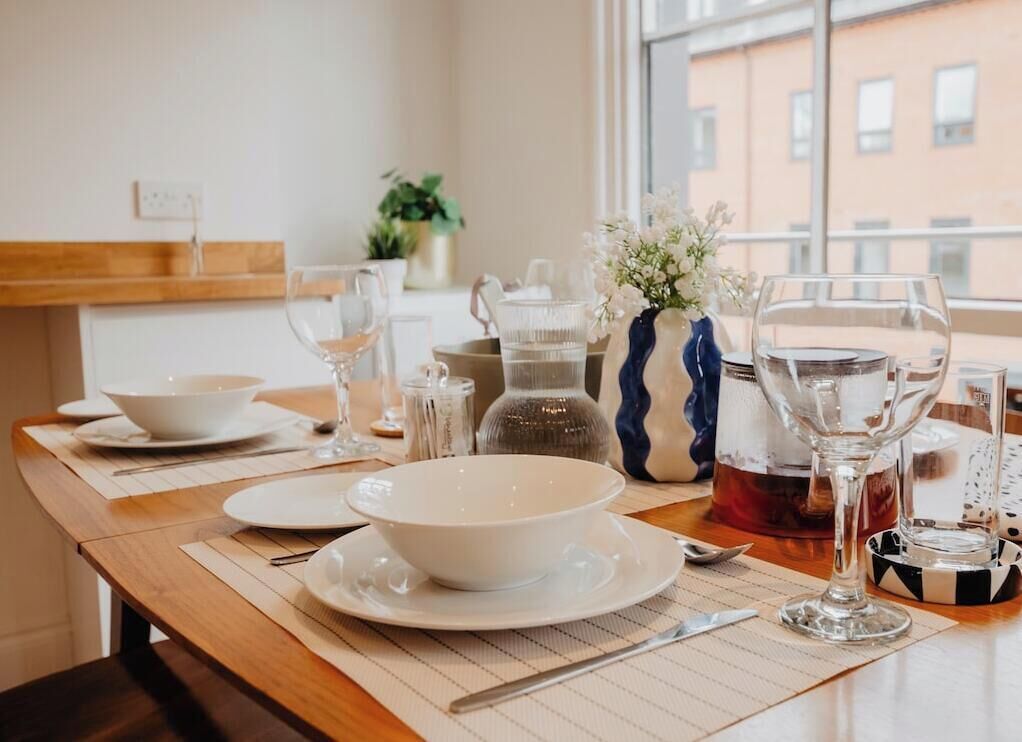 Mesa de comedor dispuesta con platos, tazones y cubiertos blancos, con un jarrón de flores y una tetera sobre la mesa.
