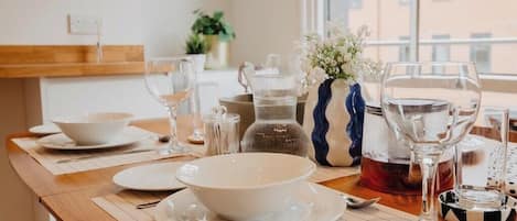 Mesa de comedor dispuesta con platos, tazones y cubiertos blancos, con un jarrón de flores y una tetera sobre la mesa.